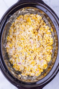 Creamy corn mixture with bits of ham in a black slow cooker on a white surface.