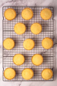 Fourteen plain cupcakes cooling on a metal rack over a marble surface.