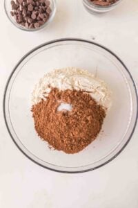 A glass bowl with flour, cocoa powder, and salt, with chocolate chips nearby.