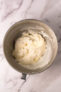 A metal mixing bowl with whipped cream cheese frosting on a marble surface.