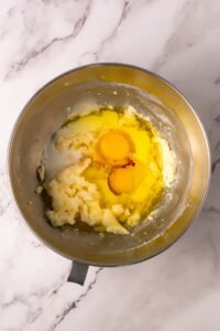 Metal bowl with creamed butter, sugar, two eggs, and vanilla on a marble surface.