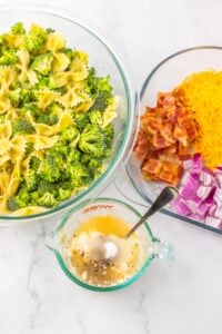 Bowtie pasta and broccoli, bacon, cheese, onions, and dressing in glass bowls.