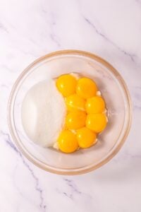A glass bowl with egg yolks and sugar on a white marble surface.