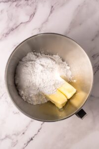 Butter and powdered sugar in a metal mixing bowl on a marble countertop.