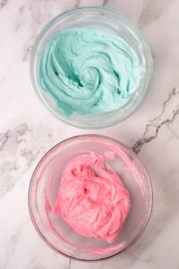 Two glass bowls with blue and pink frosting on a marble surface.