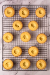 Twelve plain cupcakes with hollow centers on a cooling rack, marble background.
