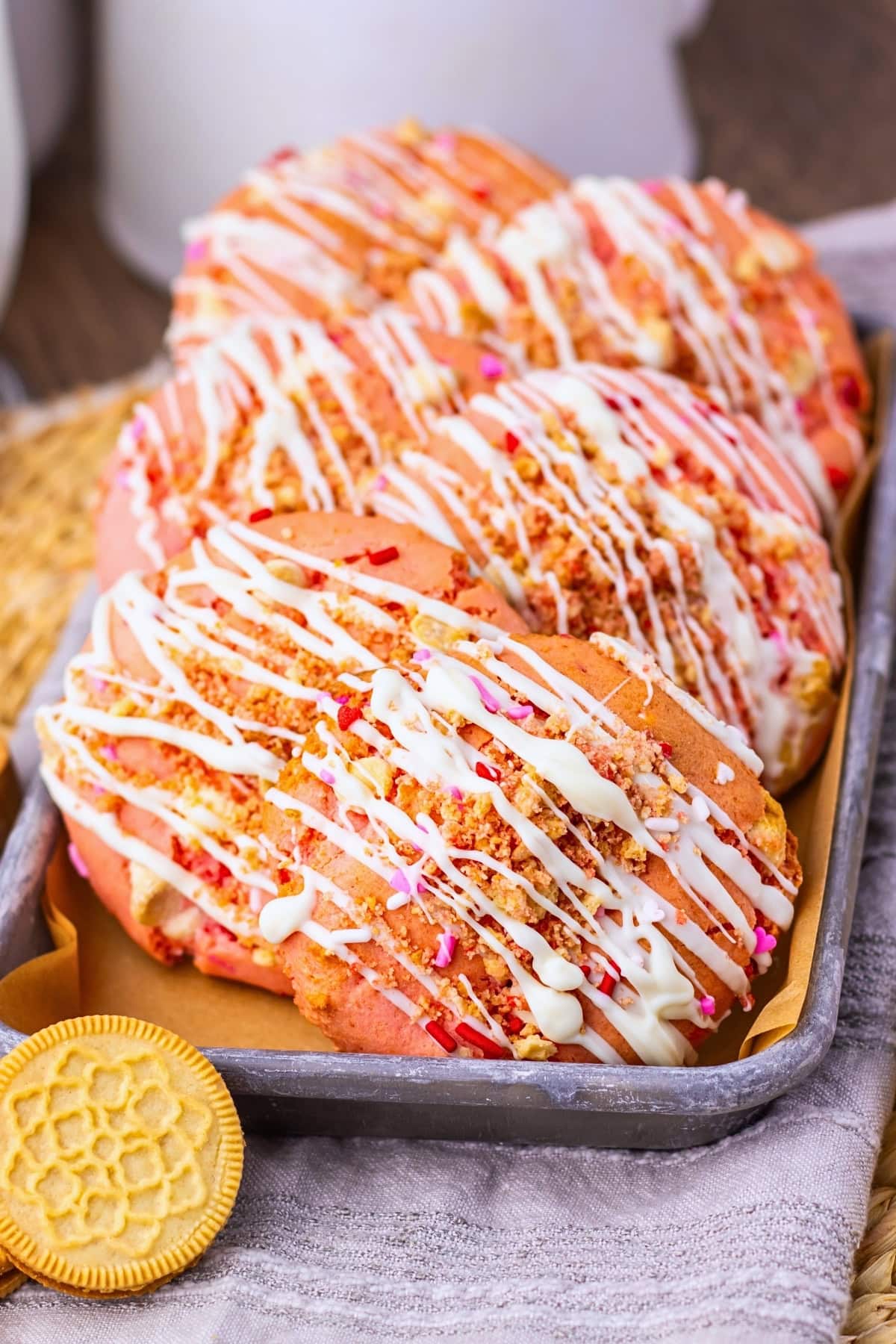 Six pink cookies with white drizzle and sprinkles on a tray next to a golden cookie.