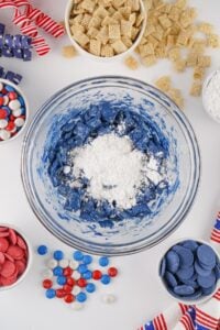 A bowl of blue snack mix with powdered sugar, surrounded by candies and cereal.