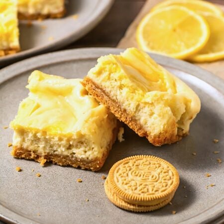 Two lemon cheesecake bars and a golden Oreo cookie on a gray plate.
