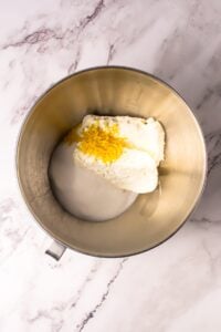 Cream cheese, sugar, and lemon zest in a metal mixing bowl on a marble surface.