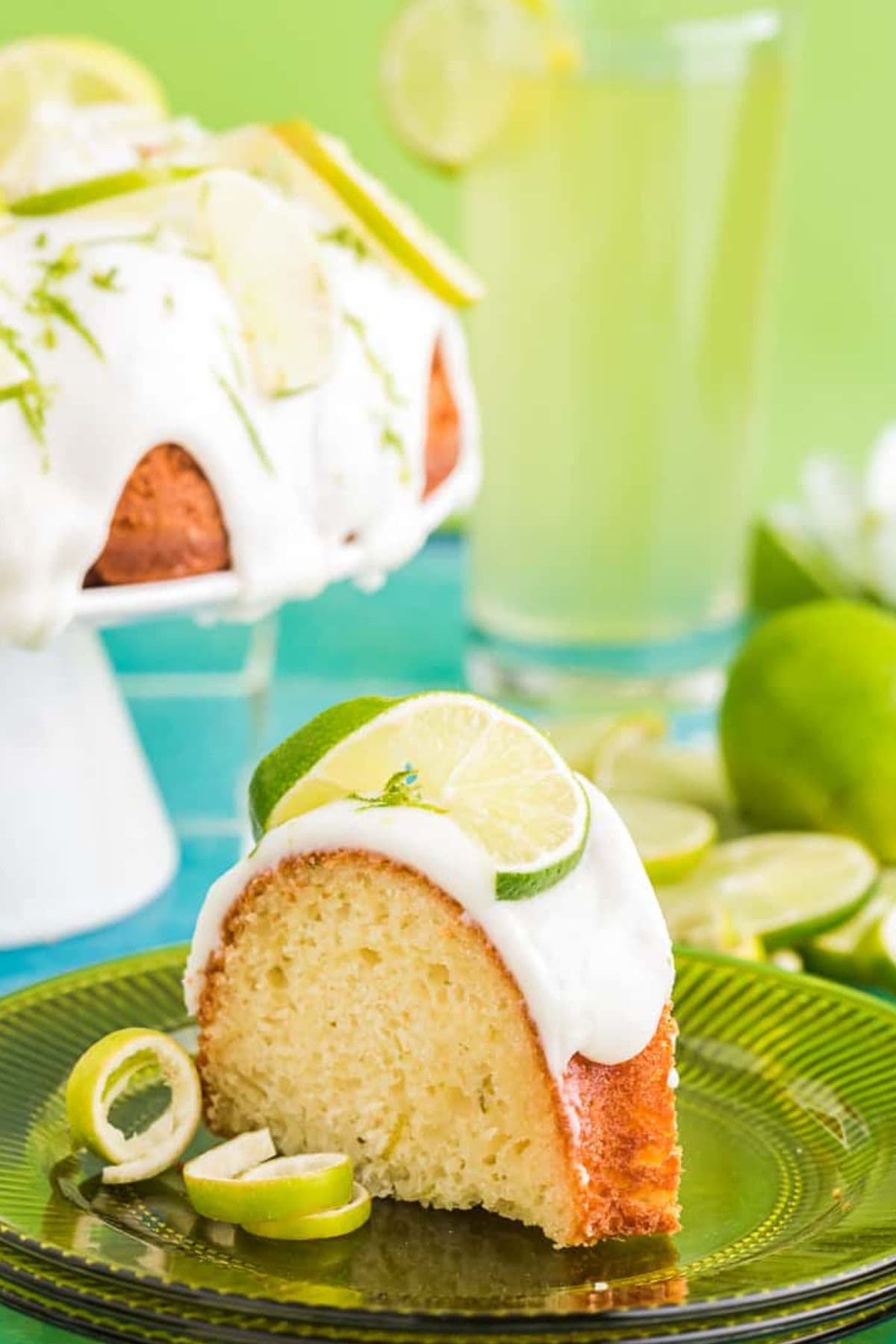Slice of lime bundt cake with white icing, lime slices, and a drink in the background.