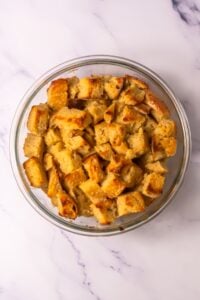 A glass bowl filled with golden-brown bread cubes on a marble surface.
