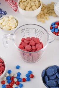 Measuring cup with red candy melts, surrounded by bowls of snacks and candies.
