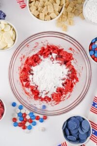 A bowl of red snack mix topped with powdered sugar, surrounded by candy and cereal.