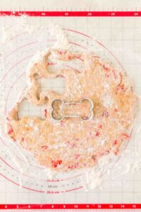 Bone-shaped cookie cutter on rolled-out dough with red bits on a floured mat.