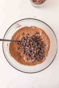 A bowl of dry brownie mix with chocolate chips and a spoon on a white surface.