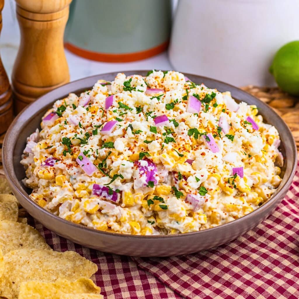 A bowl of creamy corn salad with red onions and herbs on a checkered cloth.