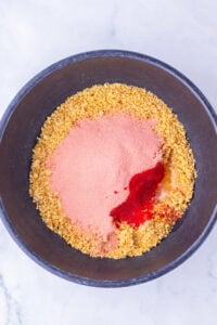 A black bowl with breadcrumbs, pink powder, and red seasoning on a white surface.