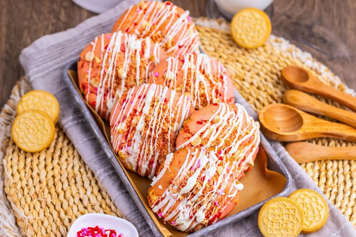 Six pink cookies drizzled with white icing and sprinkles on a tray with cookies.