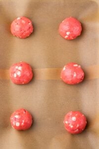 Six pink cookie dough balls with white chips on parchment paper, ready to bake.