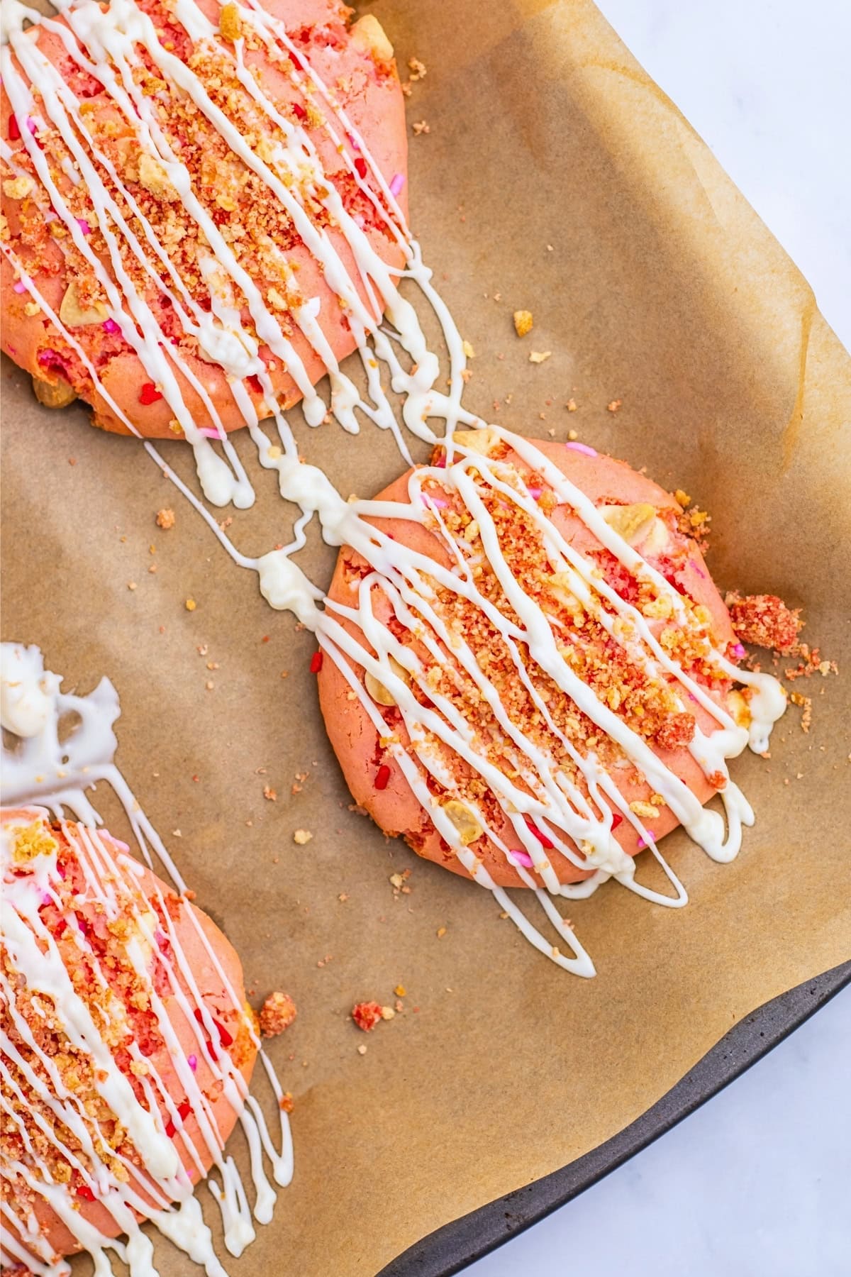 Three pink cookies with white drizzle and crumbs on parchment paper.