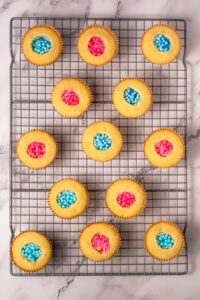 Vanilla cupcakes topped with pink and blue sprinkles on a cooling rack.