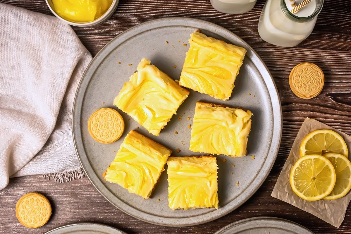 Five lemon bars on a plate with cookies, lemon slices, and bottles of milk nearby.