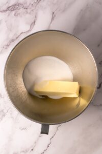 A mixing bowl with a stick of butter and granulated sugar on a marble surface.