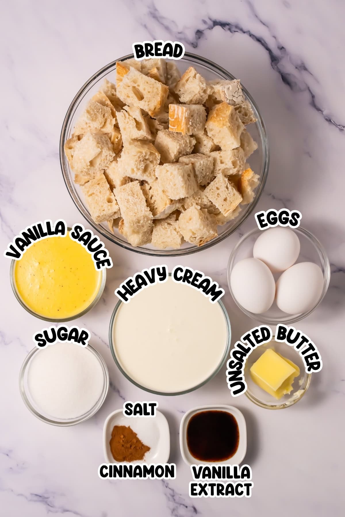 Top-down view of bread pudding ingredients in bowls, labeled on a marble surface.