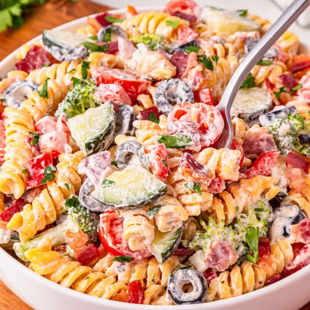 A bowl of creamy pasta salad with veggies, olives, and herbs, being served.
