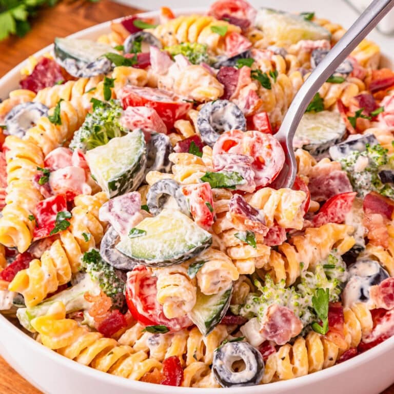 A bowl of creamy pasta salad with veggies, olives, and herbs, being served.