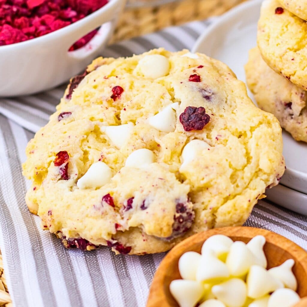 A cookie with white chocolate chips and red berry pieces on a striped cloth.