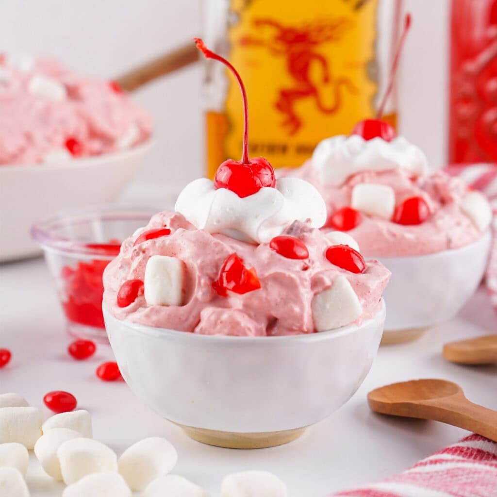 Pink dessert topped with cherries, marshmallows, and whipped cream in a white bowl.