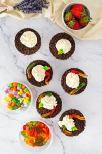 Six dirt cup desserts with gummy worms and whipped cream, plus candies and strawberries.