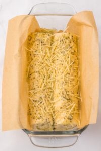 Unbaked bread dough topped with shredded cheese and herbs in a parchment-lined pan.