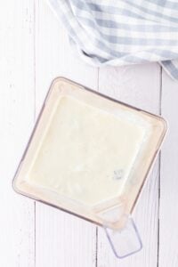 Blender with creamy white mixture on a white wooden surface, next to a cloth.