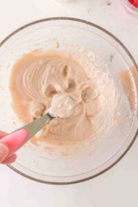 A hand holds a spoon of batter over a glass bowl filled with creamy batter.
