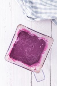 Top view of a blender with thick purple smoothie on a white wooden surface.