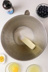 Stand mixer bowl with butter and sugar, surrounded by eggs, lemon, blueberries, vanilla.