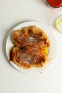 Two raw chicken breasts on a white plate, sprinkled with various spices.