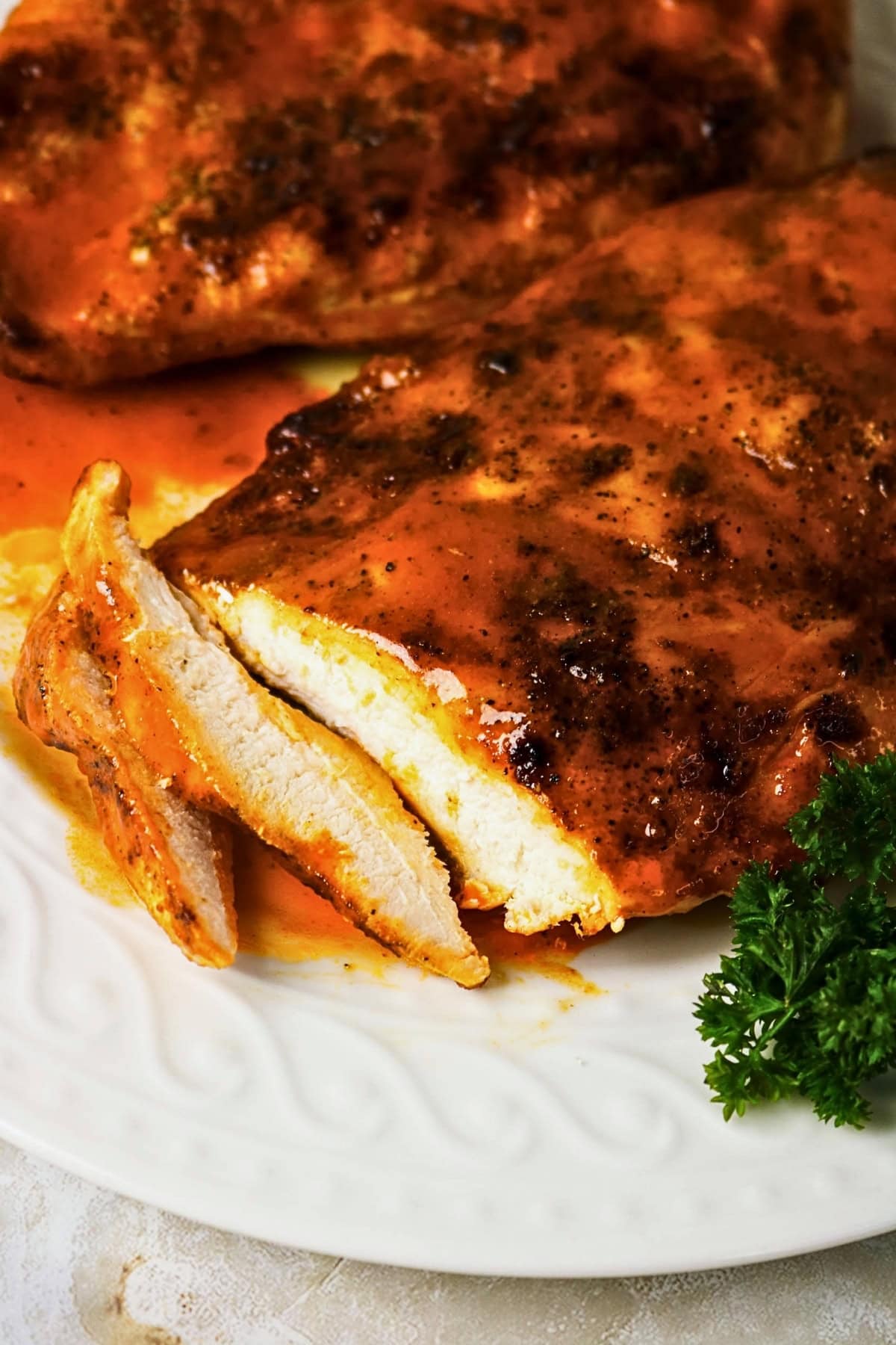 Sliced baked chicken breast with sauce on a white plate, garnished with parsley.