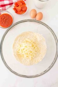 Glass bowl with flour and shredded cheese, eggs, sauce, and pepperoni nearby.