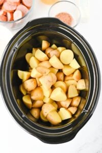 Quartered potatoes in a slow cooker, with sliced sausage in a bowl nearby.