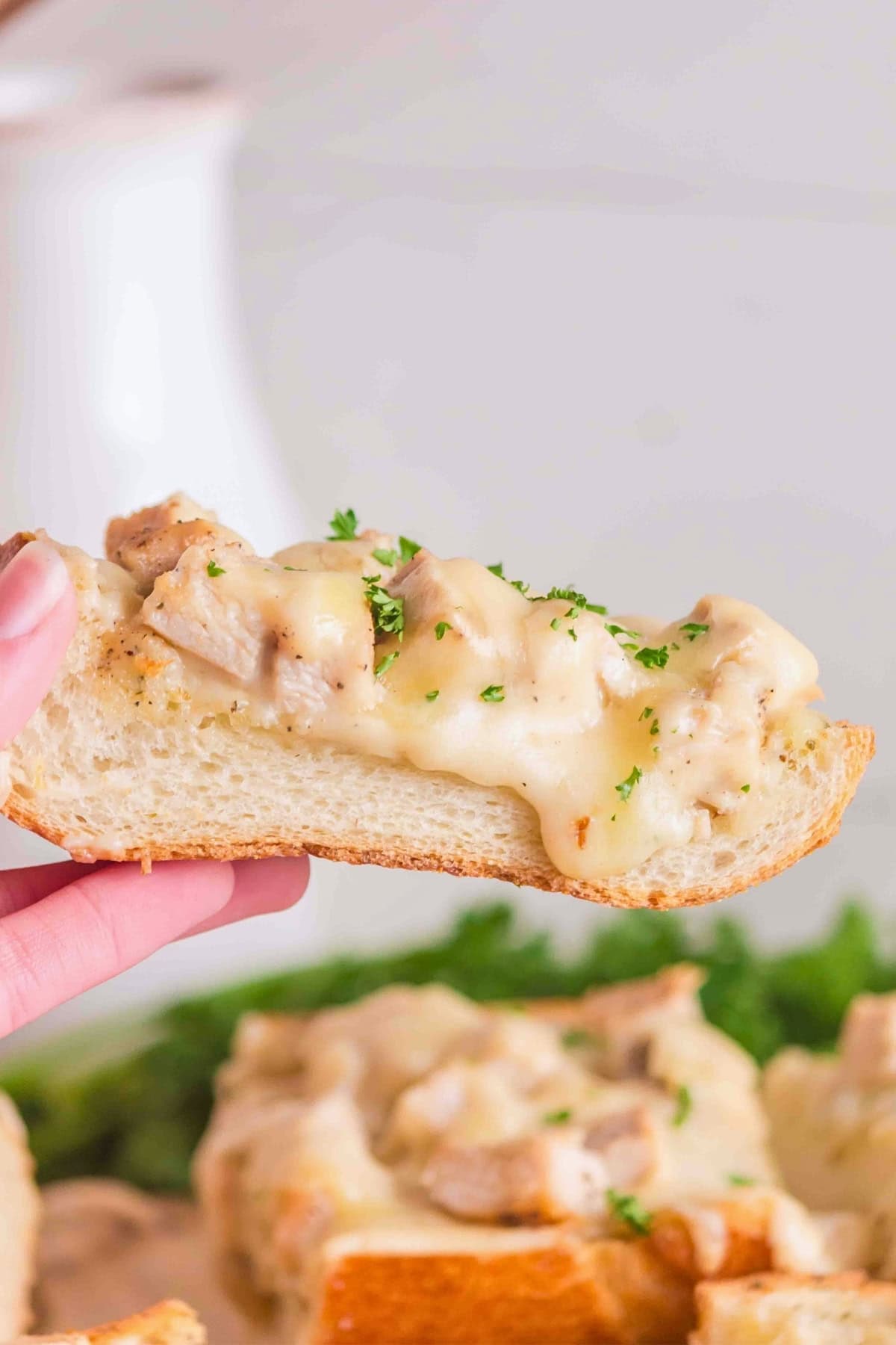 A hand holds a slice of bread topped with creamy chicken and herbs.