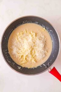 Shredded cheese melting into a creamy sauce in a black pan with a red handle.
