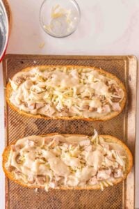 Two sandwich halves topped with diced chicken, cheese, and sauce on a baking sheet.