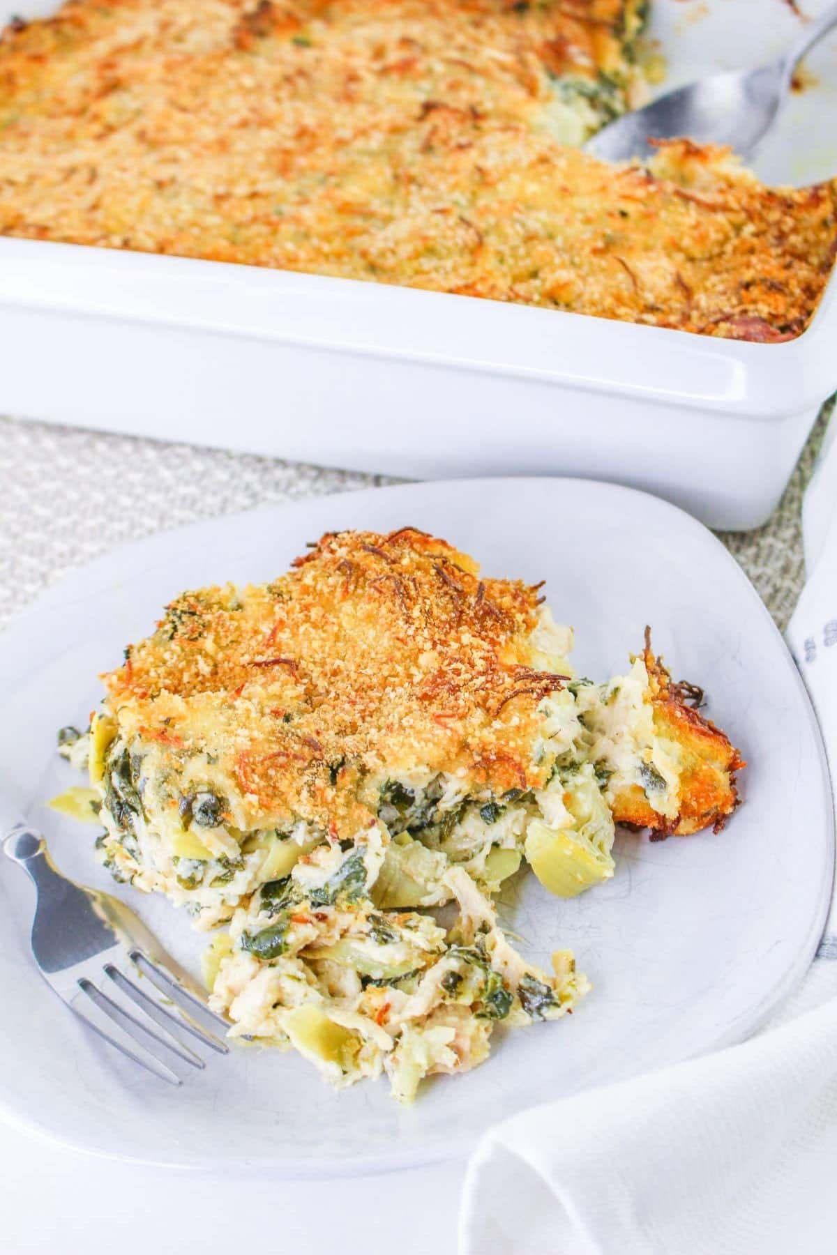 A slice of baked spinach artichoke chicken casserole on a white plate with a fork.