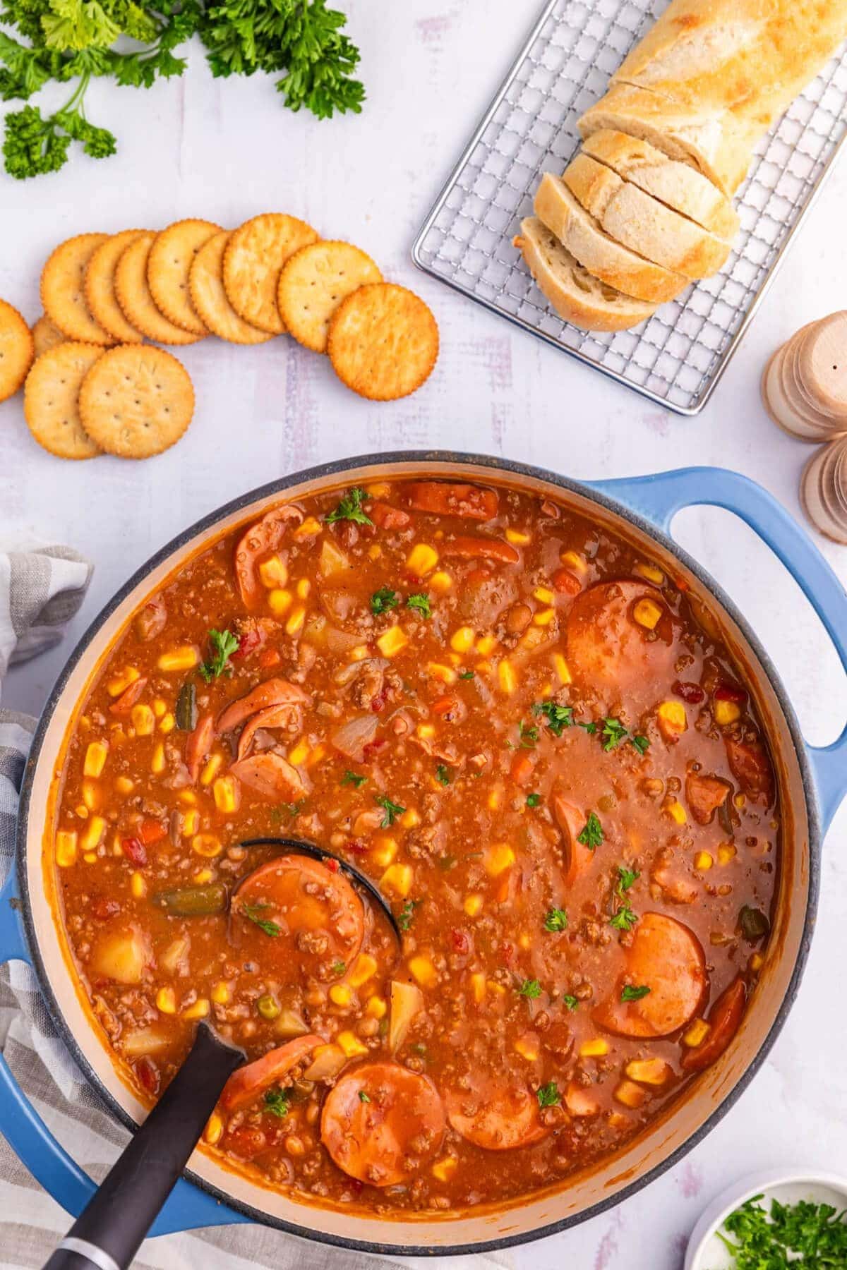 A pot of kielbasa sausage soup beside crackers and sliced bread on a cooling rack.