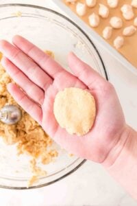 A hand holding a round, flattened ball of cookie dough above a mixing bowl.