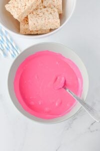 A bowl of pink melted candy with a spoon, next to rice crispy treats.